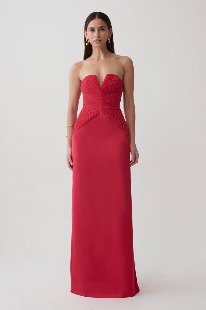 A woman modeling a strapless red floor-length gown with a sweetheart neckline, ruched bodice, and gathered waist details, photographed against a plain light gray studio background.