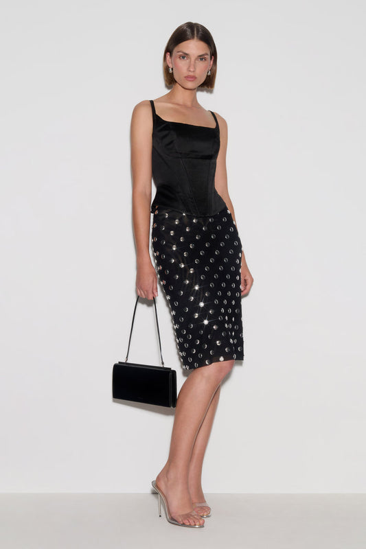 A model wearing a black satin camisole top and a black studded pencil skirt, holding a black velvet clutch bag, posed against a plain white studio background.