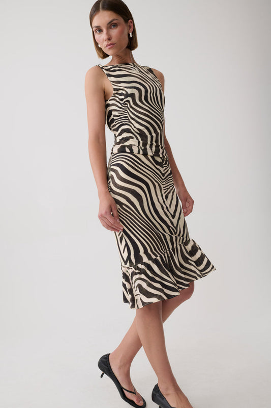 A woman modeling a sleeveless halter-neck midi dress with a bold black and beige zebra-like wavy stripe pattern, paired with black heels, against a plain light gray studio background.