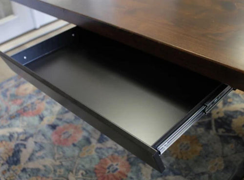 A Standard Desk Add-On by James+James featuring a wooden top with an open black metal drawer beneath it. The drawer is empty, and the desk stands on a colorful, patterned rug with floral designs. In the background, part of a white door and a light-colored floor are visible.